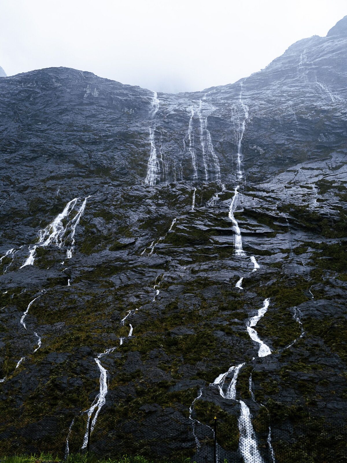 An einem nebligen Tag stürzt Wasser eine steile, felsige Bergwand hinab und bildet mehrere schmale Ströme über dunklem, strukturiertem Stein. (KI-generierte Beschreibung)