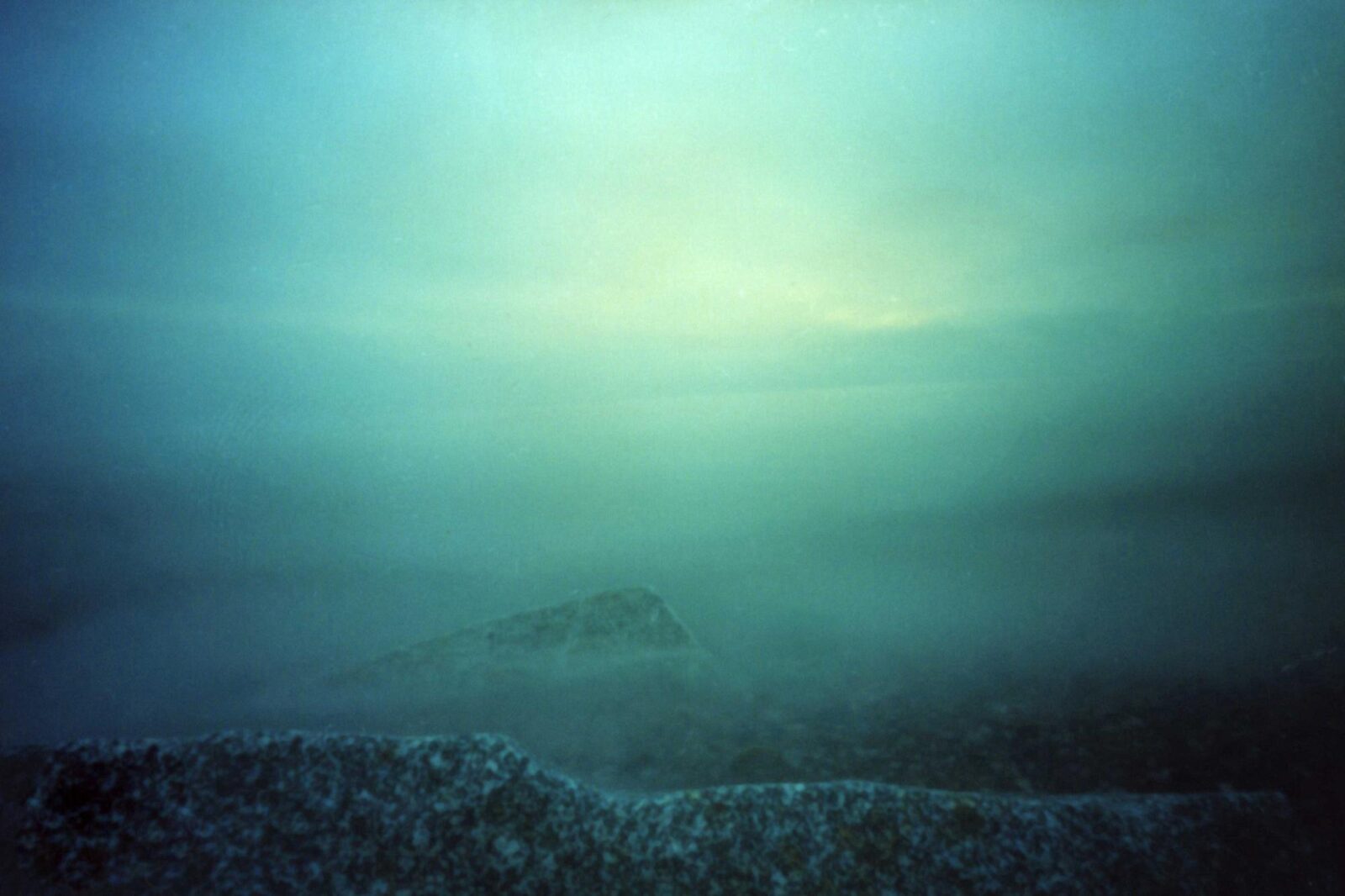 Neblige Meereslandschaft mit nebligem Horizont und undeutlichen Umrissen entfernter Felsen unter einem grünlichen Himmel. (KI-generierte Beschreibung)