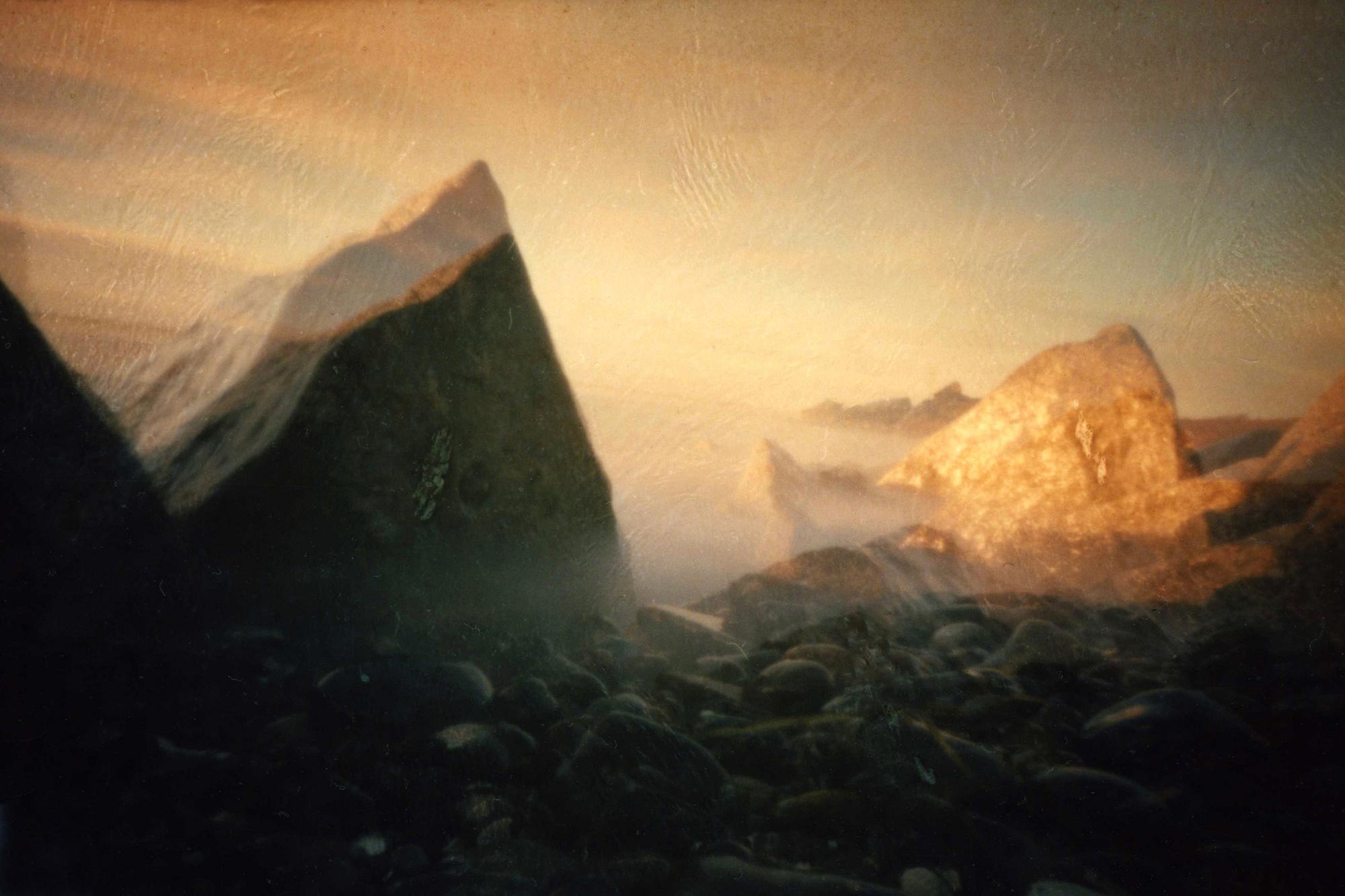 Abstraktes Bild, das eine felsige Landschaft mit einem warmen, ätherischen Glanz darstellt. Große, scharfe Felsformationen dominieren den Vordergrund, wobei eine neblige Atmosphäre entweder Wasser- oder Luftbewegungen suggeriert. (KI-generierte Beschreibung)