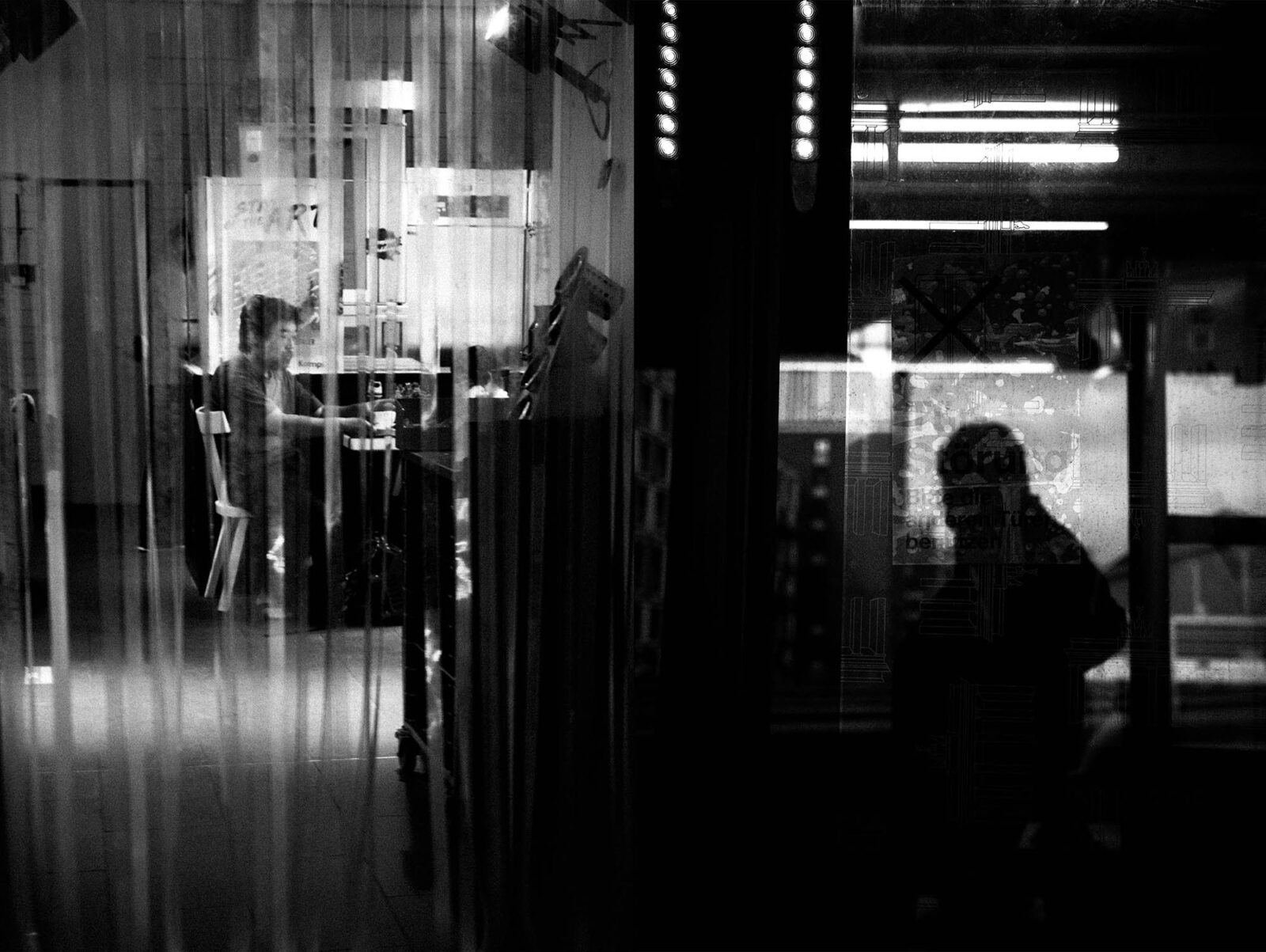 Schwarzweißbild einer Person, die hinter einem durchsichtigen Vorhang an einem Schreibtisch arbeitet, während sich in einem Fenster eine weitere Silhouette spiegelt. (KI-generierte Beschreibung)