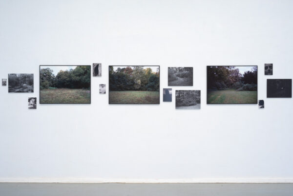 Eine Reihe von gerahmten Fotografien, die Landschaften im Freien zeigen, und kleinere Schwarz-Weiß-Porträts sind auf einer weißen Galeriewand ausgestellt. (KI-generierte Beschreibung)