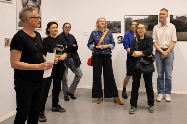Eine Gruppe Erwachsener steht in einer Kunstgalerie zusammen, einige lächeln, während eine Person Papiere hält und zu der Gruppe zu sprechen scheint. An den Wänden hinter ihnen sind gerahmte Fotografien zu sehen. (KI-generierte Beschreibung)