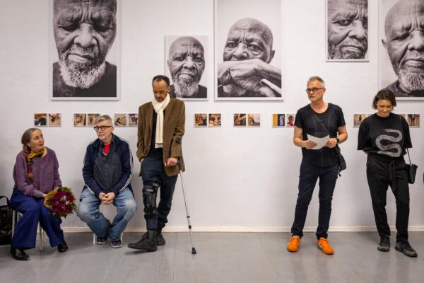 Fünf Personen stehen oder sitzen vor einer Galeriewand, an der große Schwarz-Weiß-Porträts eines älteren Mannes und kleinere Farbfotos hängen. Eine Person hält einen Stock, eine andere hält Papiere in der Hand. (KI-generierte Beschreibung)