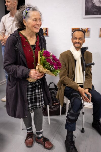 Eine ältere Frau mit einem Blumenstrauß in der Hand steht lächelnd neben einem sitzenden Mann mit einer Beinschiene und einem Stock; andere Personen sind teilweise im Hintergrund zu sehen. (KI-generierte Beschreibung)