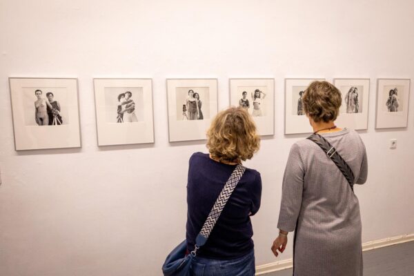 Zwei Frauen stehen vor einer weißen Wand und betrachten eine Reihe von gerahmten Schwarz-Weiß-Fotografien in einer Kunstgalerie. (KI-generierte Beschreibung)