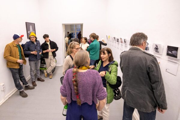 Eine Gruppe von Menschen unterhält sich und betrachtet Fotografien, die an den weißen Wänden eines hell erleuchteten Galerieraums ausgestellt sind. (KI-generierte Beschreibung)