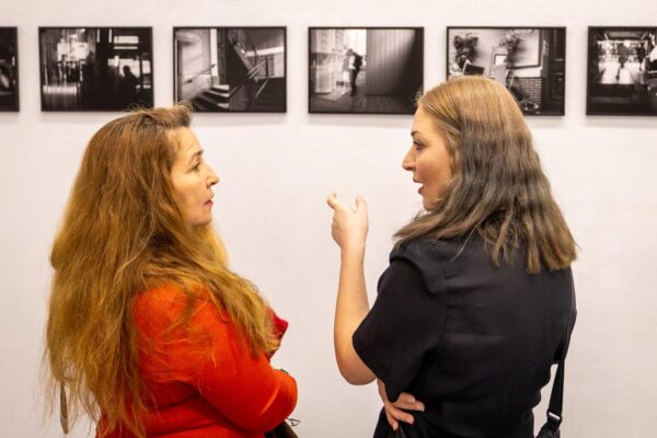 Zwei Frauen stehen sich gegenüber und unterhalten sich vor einer Wand, auf der eine Reihe von Schwarz-Weiß-Fotos zu sehen ist. (KI-generierte Beschreibung)
