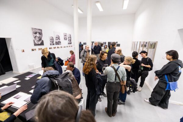 Menschen versammeln sich in einer weißen Kunstgalerie, betrachten Fotos an den Wänden und unterhalten sich an Tischen mit Büchern und Papieren. (KI-generierte Beschreibung)