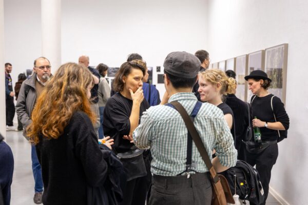 Eine Gruppe von Menschen unterhält sich und betrachtet gerahmte Kunstwerke an den Wänden einer Indoor-Galerieausstellung. (KI-generierte Beschreibung)