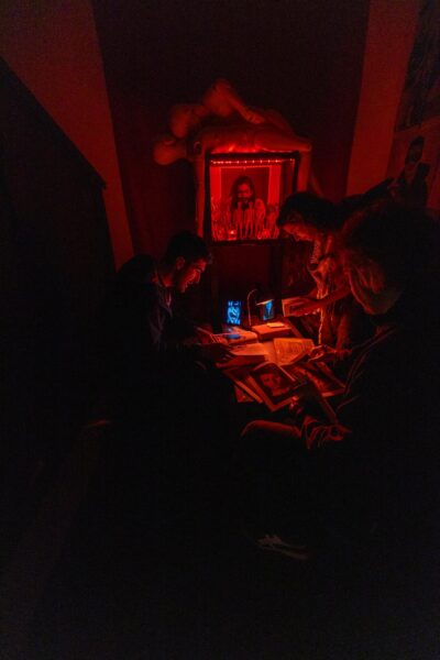 Vier Personen sitzen in einem schwach beleuchteten roten Raum um einen Tisch herum und prüfen Bücher und Papiere unter rotem Licht, während im Hintergrund ein gerahmtes Foto und eine Schaufensterpuppe zu sehen sind. (KI-generierte Beschreibung)