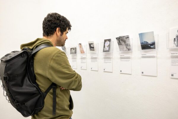 Eine Person mit schwarzem Rucksack und olivgrünem Kapuzenpulli betrachtet eine Ausstellung von Fotos und Texttafeln an einer weißen Wand. (KI-generierte Beschreibung)
