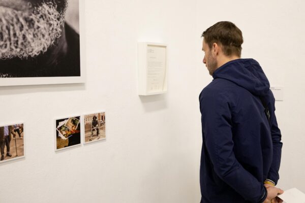 Ein Mann in einer blauen Jacke steht vor einer weißen Wand und betrachtet Fotos und ein gerahmtes Dokument in einer Kunstgalerie oder Ausstellung. (KI-generierte Beschreibung)