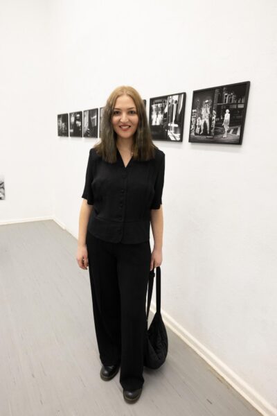 Eine schwarz gekleidete Frau steht in einer Kunstgalerie mit Schwarz-Weiß-Fotografien an der weißen Wand hinter ihr. (KI-generierte Beschreibung)